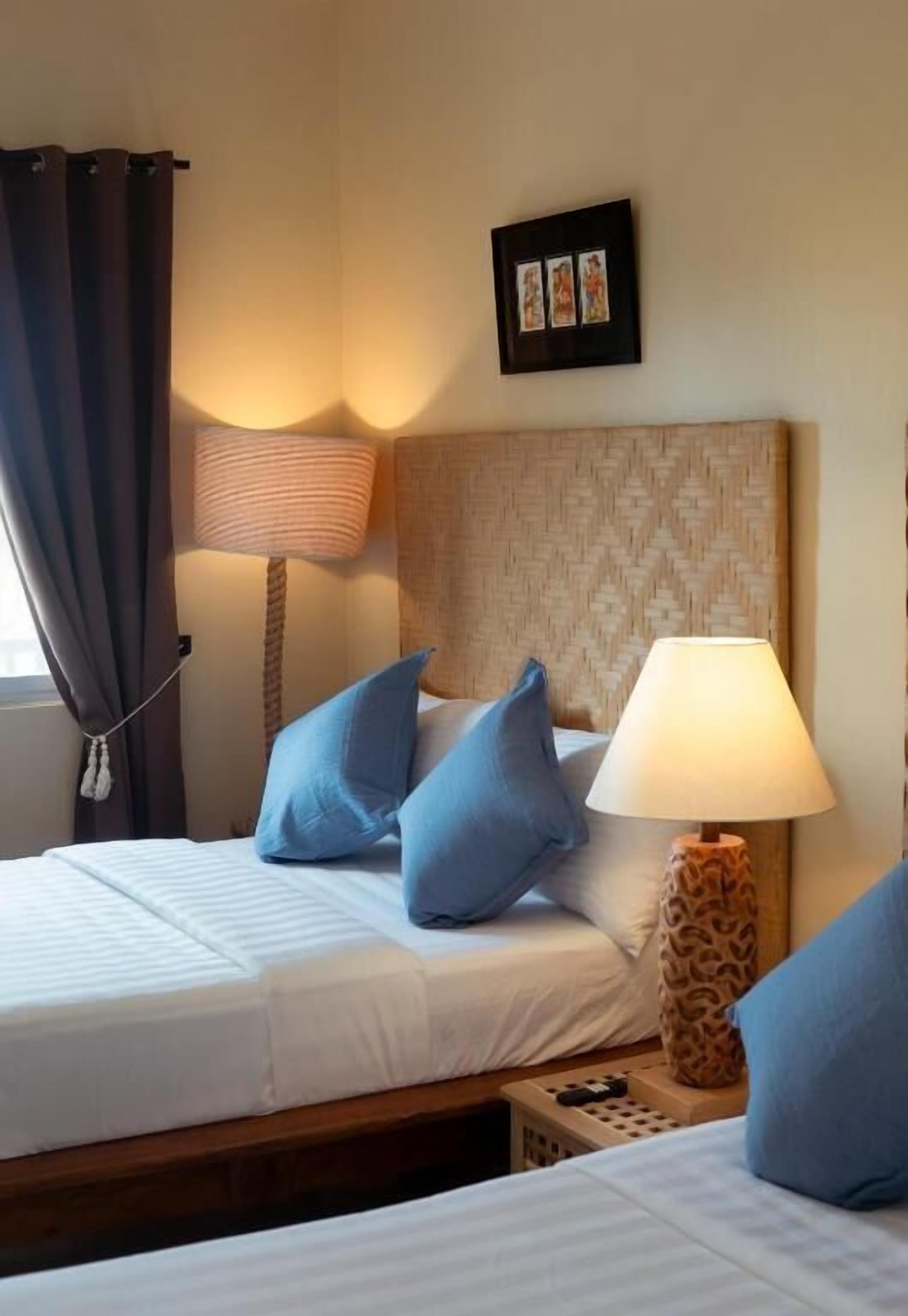 Ein Hotelzimmer mit einem Bett, zwei Kissen, einer Lampe, einem Bilderrahmen und einem Fenster mit Vorhängen.