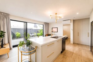 A modern kitchen with a large island, a dining area with a table and chairs, and a view of a garden through a sliding door. - Christchurch 2BR | Courtyard + Free Parking (Christchurch)