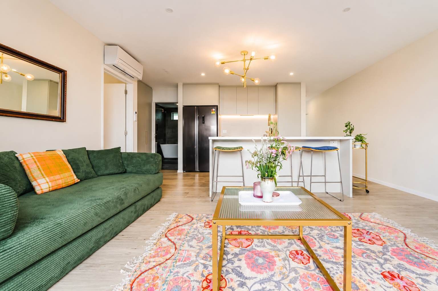 Una sala moderna con un sofá verde, una mesa de centro con un jarrón de flores y una cocina al fondo.