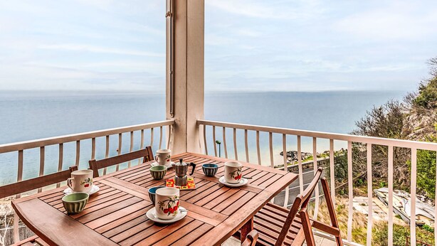 A wooden table with cups and a coffee pot on a balcony overlooking the sea.