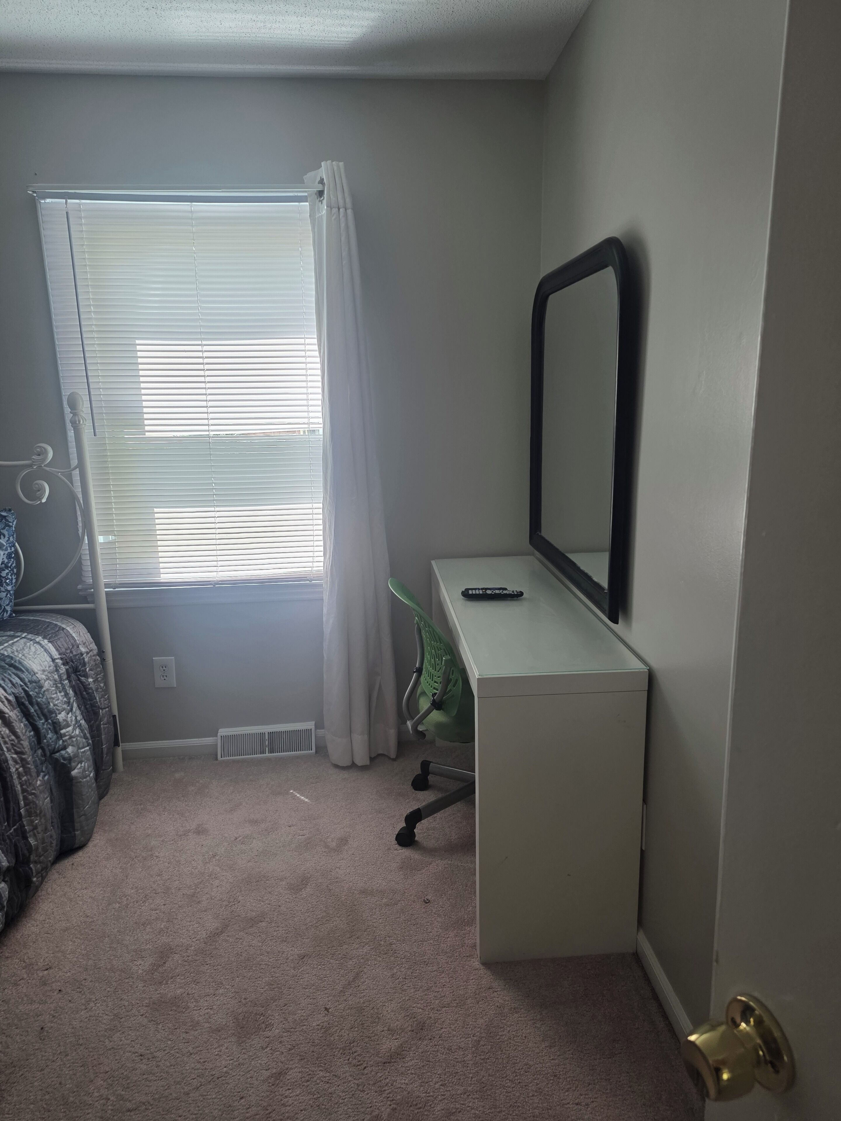 A bedroom with a bed, a desk, a chair, a window with curtains, and a door.