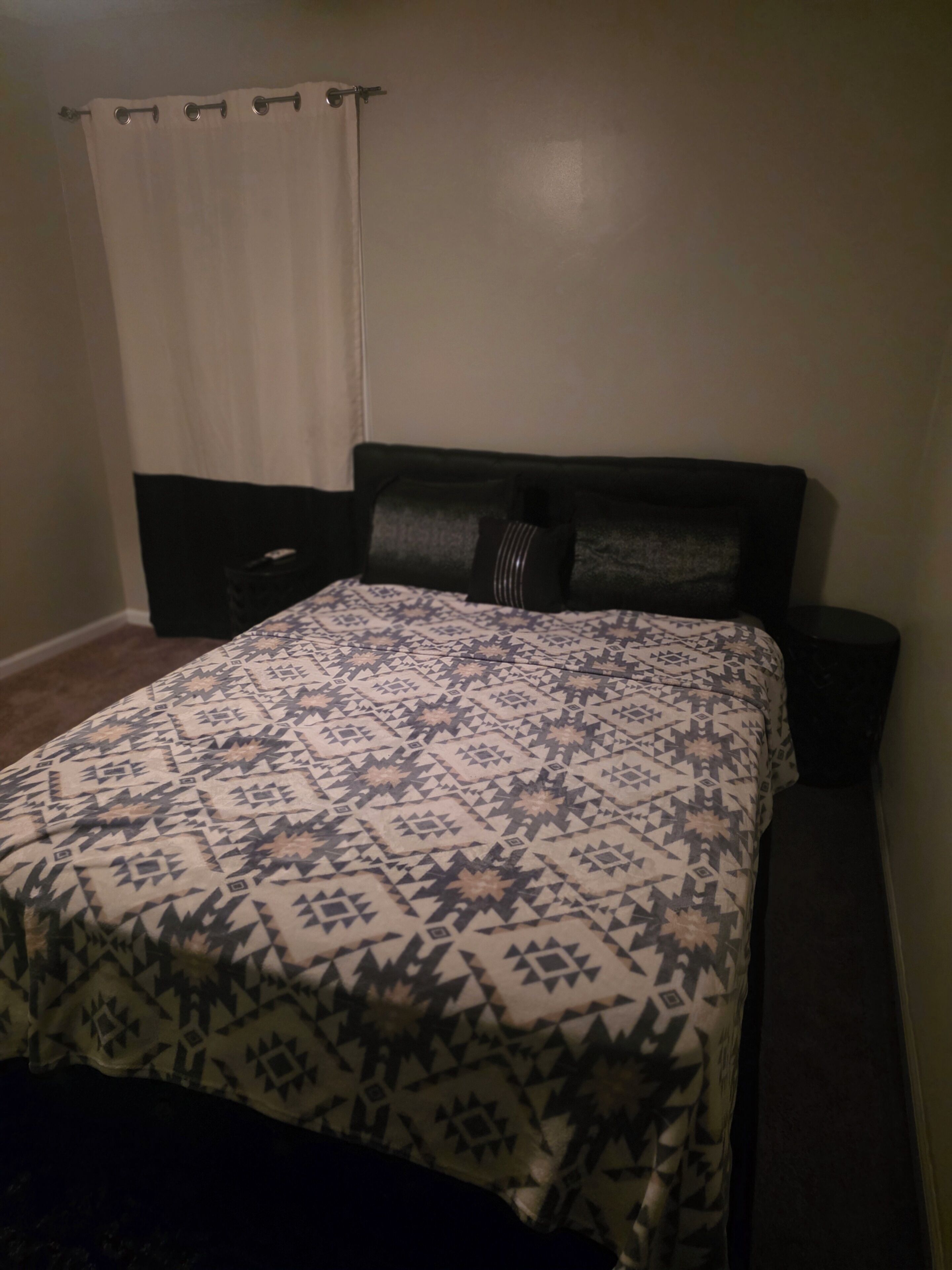 Een slaapkamer met een bed, nachtkastje, donker hoofdbord en een witte gordijn.
