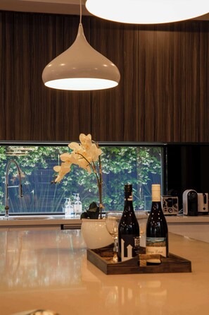 Eine moderne Küche mit Holzwand, einer weißen Pendelleuchte, einer Vase mit weißen Blumen und einer Ablage mit Weinflaschen und Gläsern.