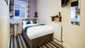 A small, neatly arranged bedroom with a bed, a bedside table, a mirror, a radiator, and a window with curtains.