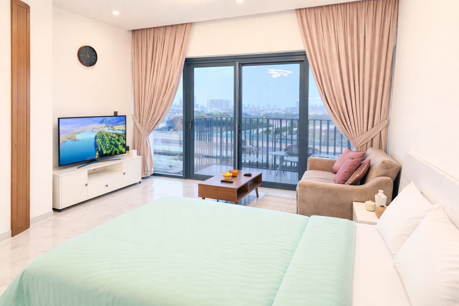 A modern bedroom with a large bed, a TV on a stand, a sofa, and a balcony with a city view.