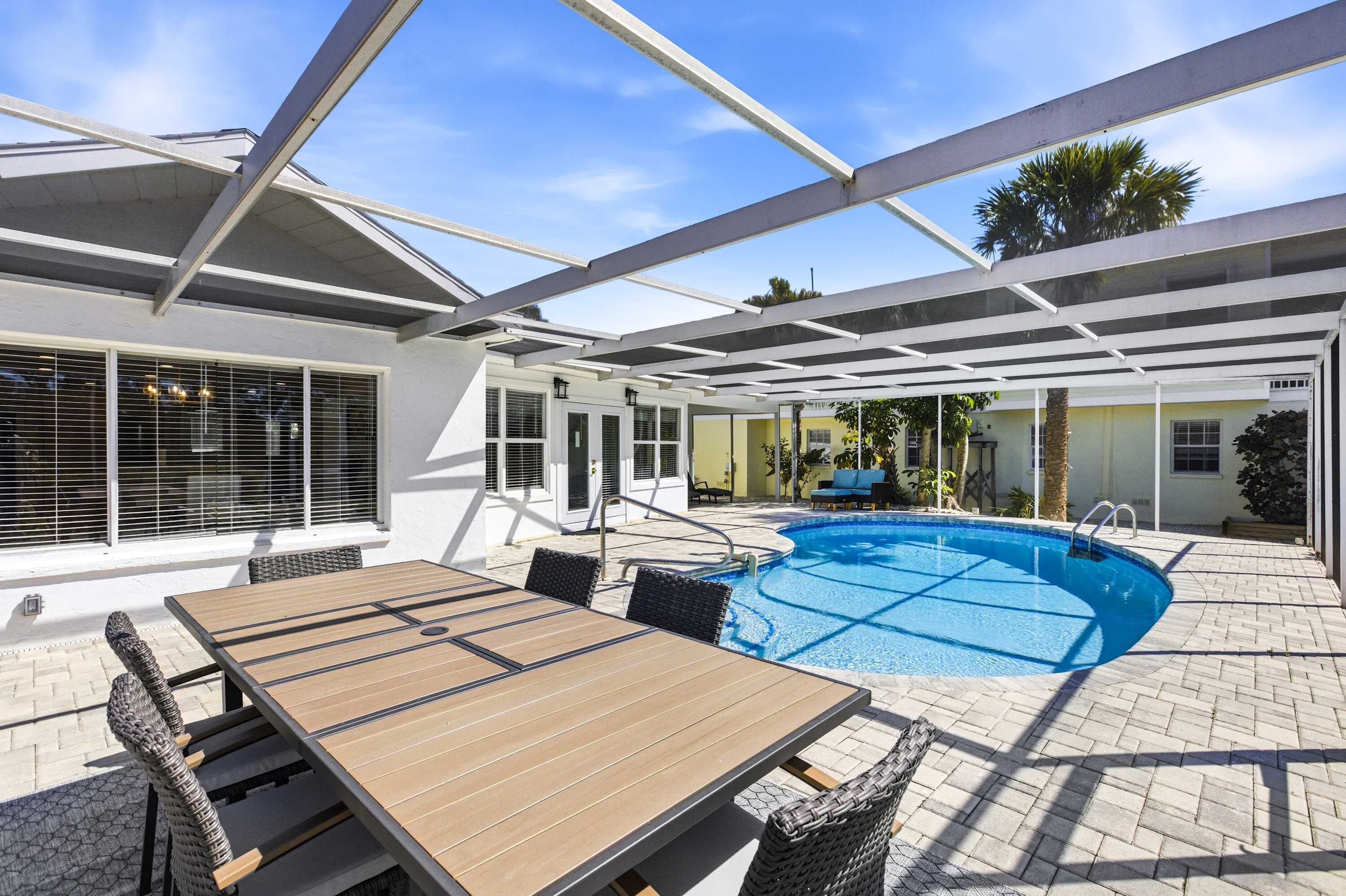 An outdoor pool area with a wooden table and chairs, a covered patio, and a clear blue sky.