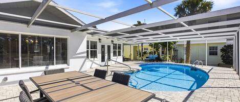 An outdoor pool area with a wooden table and chairs, a covered patio, and a clear blue sky.