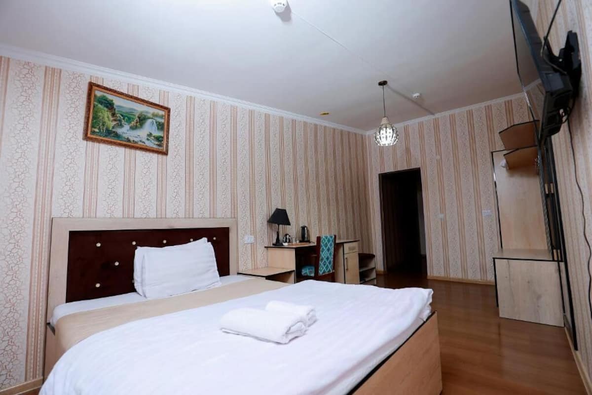 A hotel room with a large bed, a nightstand, a desk, a chair, a TV mounted on the wall, and a door leading to another room.