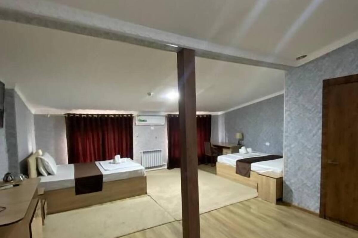 A spacious hotel room with a large bed, a desk, and a chair. There is a window with curtains, a radiator, and a door.
