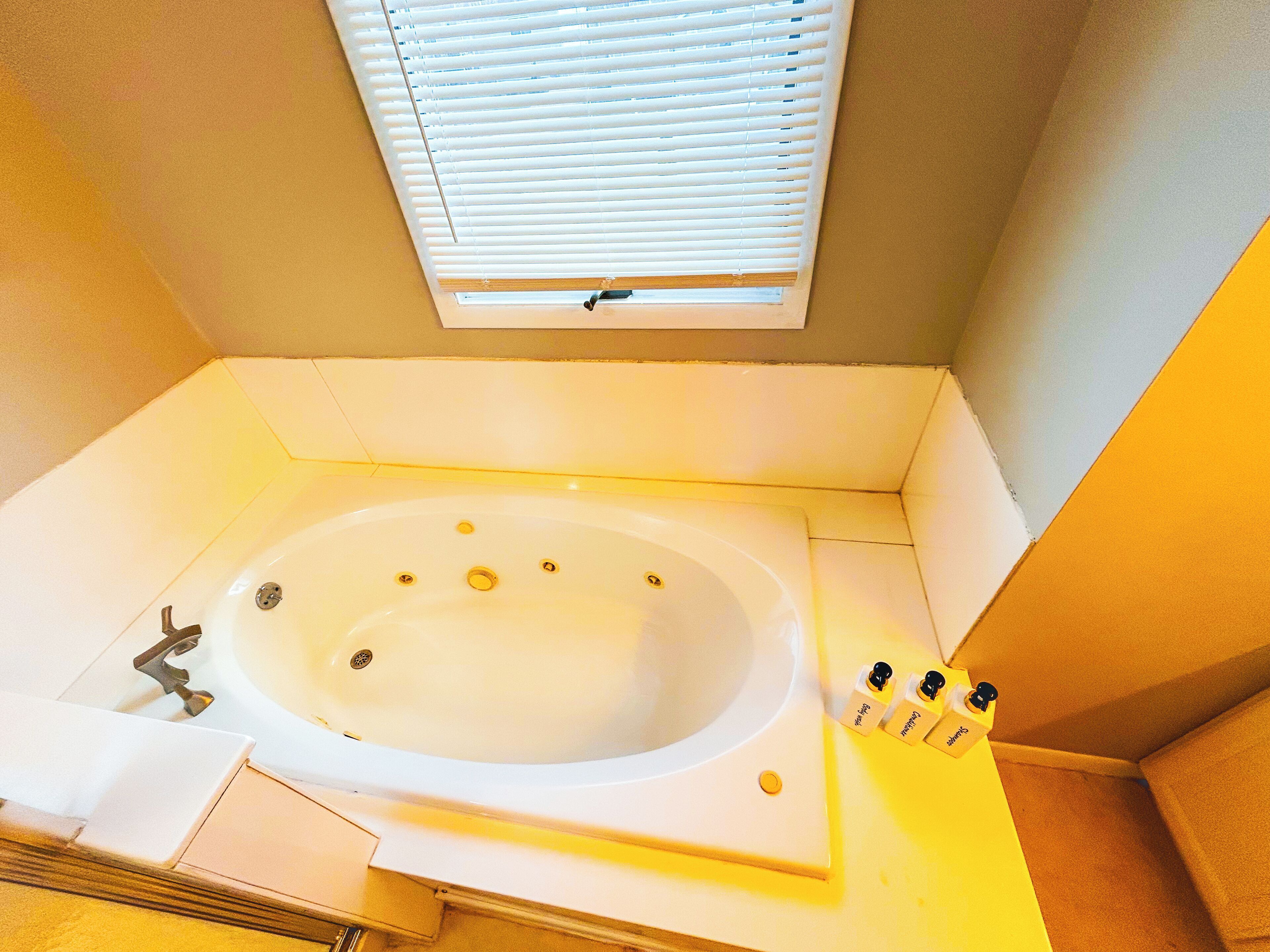 Una bañera con una ventana encima, un cabezal de ducha y un juego de artículos de aseo a un lado.