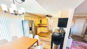 Cocina compacta con refrigerador, microondas y estufa. Hay una zona de comedor con mesa y sillas. El cuarto tiene piso de madera y un ventilador de techo.