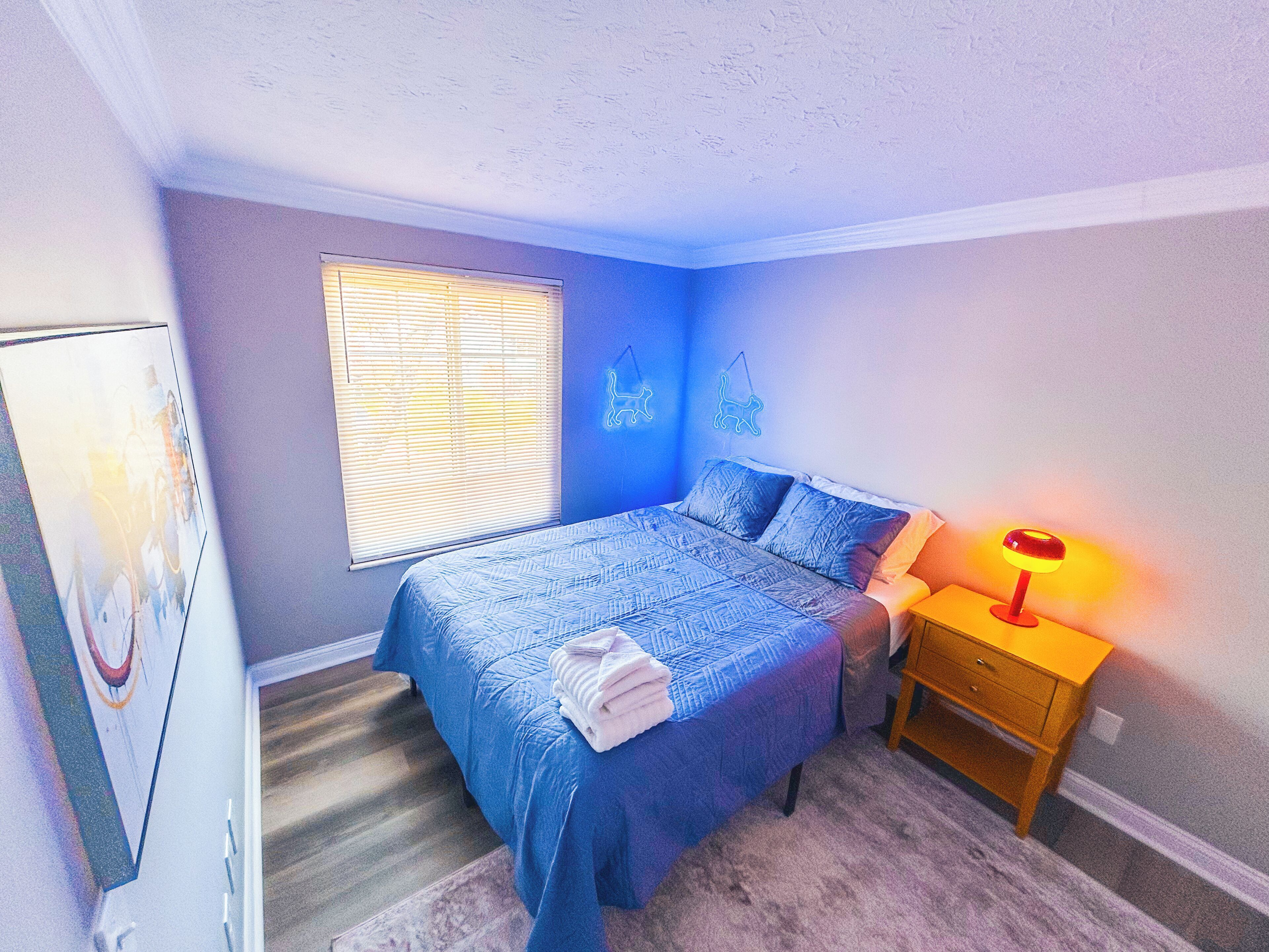 Ein Schlafzimmer mit Bett, Nachttisch, Lampe, einem Fenster mit Jalousien und einer Wanddekoration.