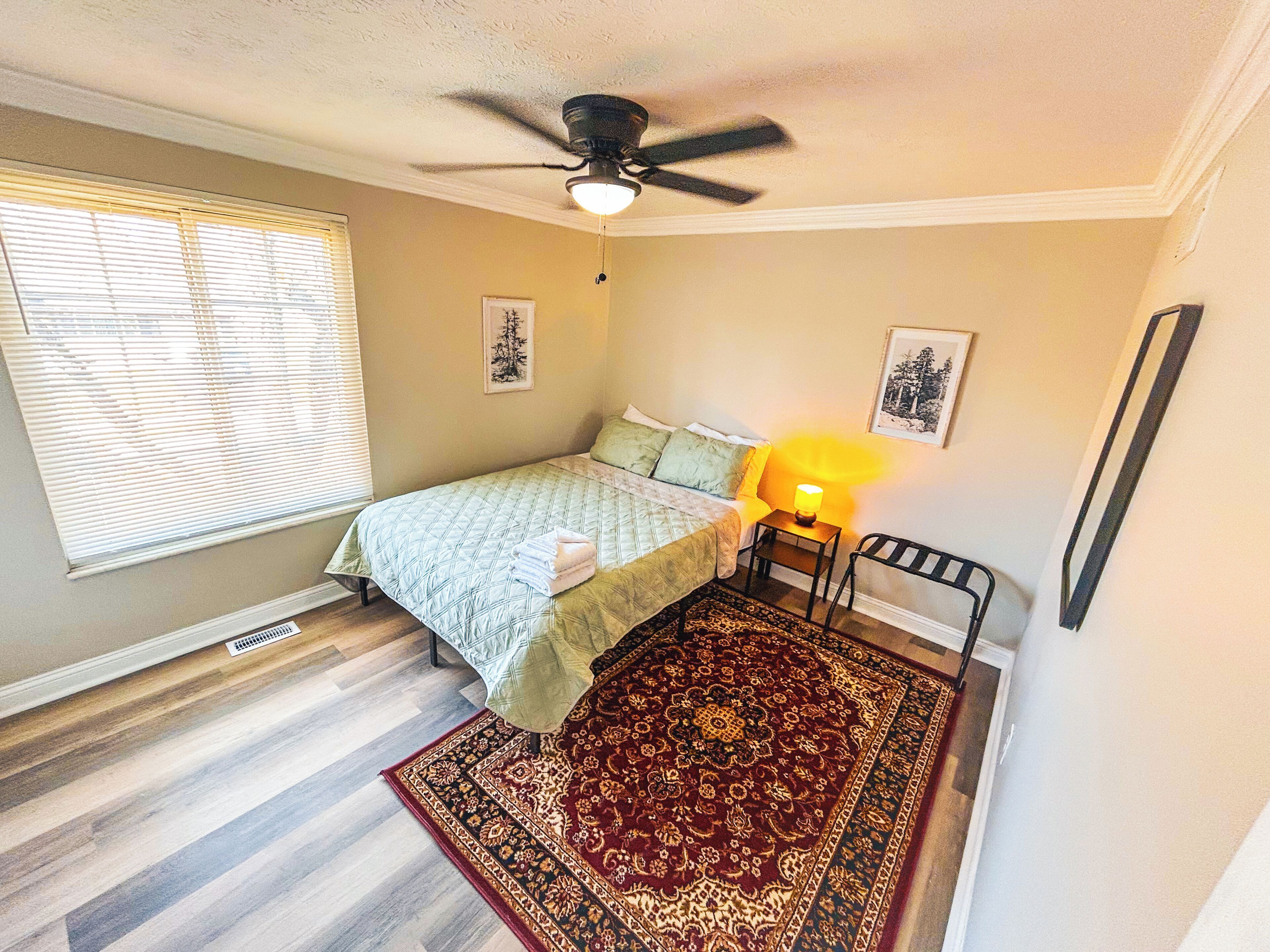 Une chambre à coucher avec un lit, un ventilateur de plafond, une lampe de chevet, un tapis et deux tableaux encadrés accrochés au mur.