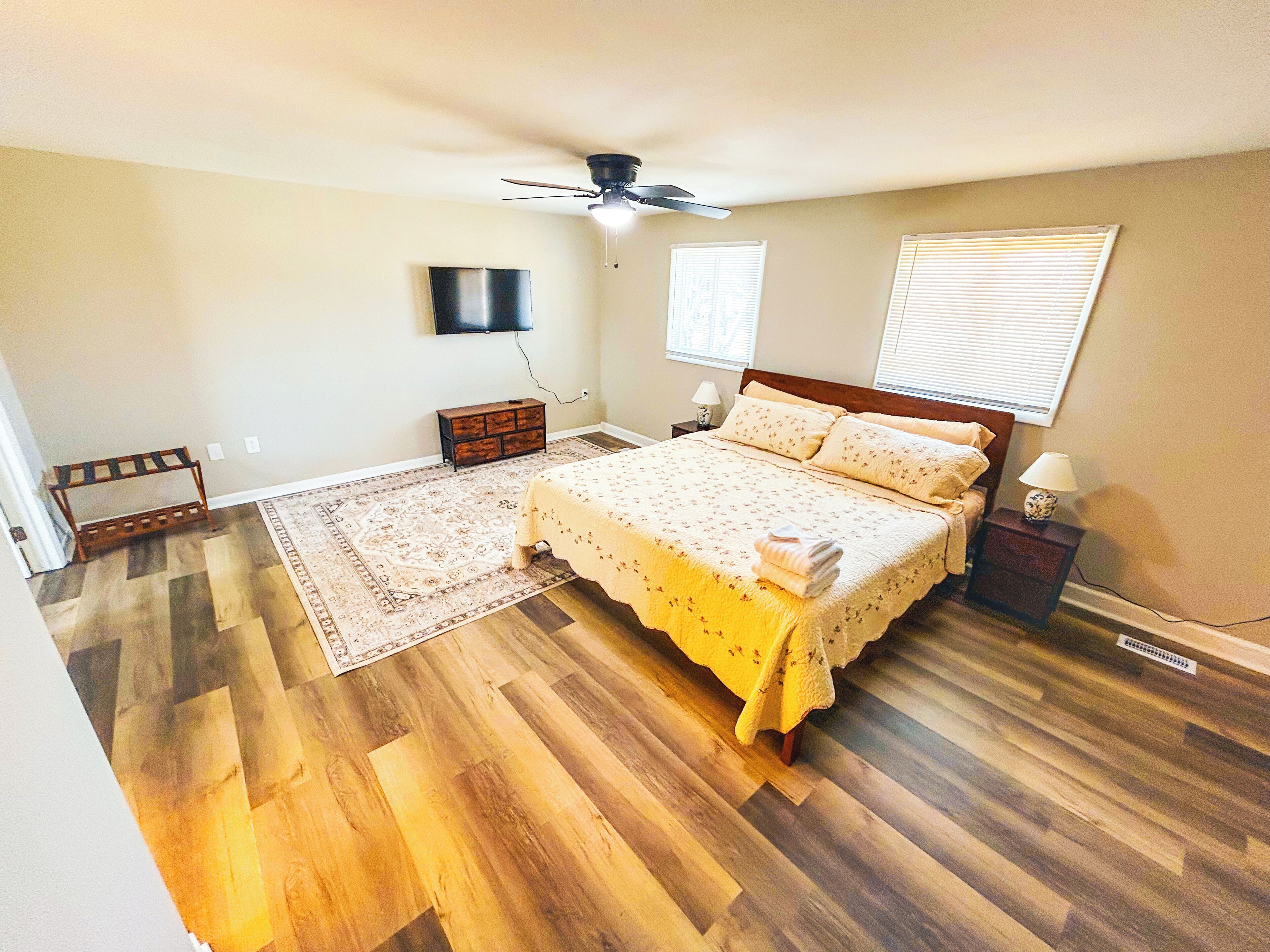 A bedroom with a bed, a ceiling fan, a TV mounted on the wall, and a window with blinds.