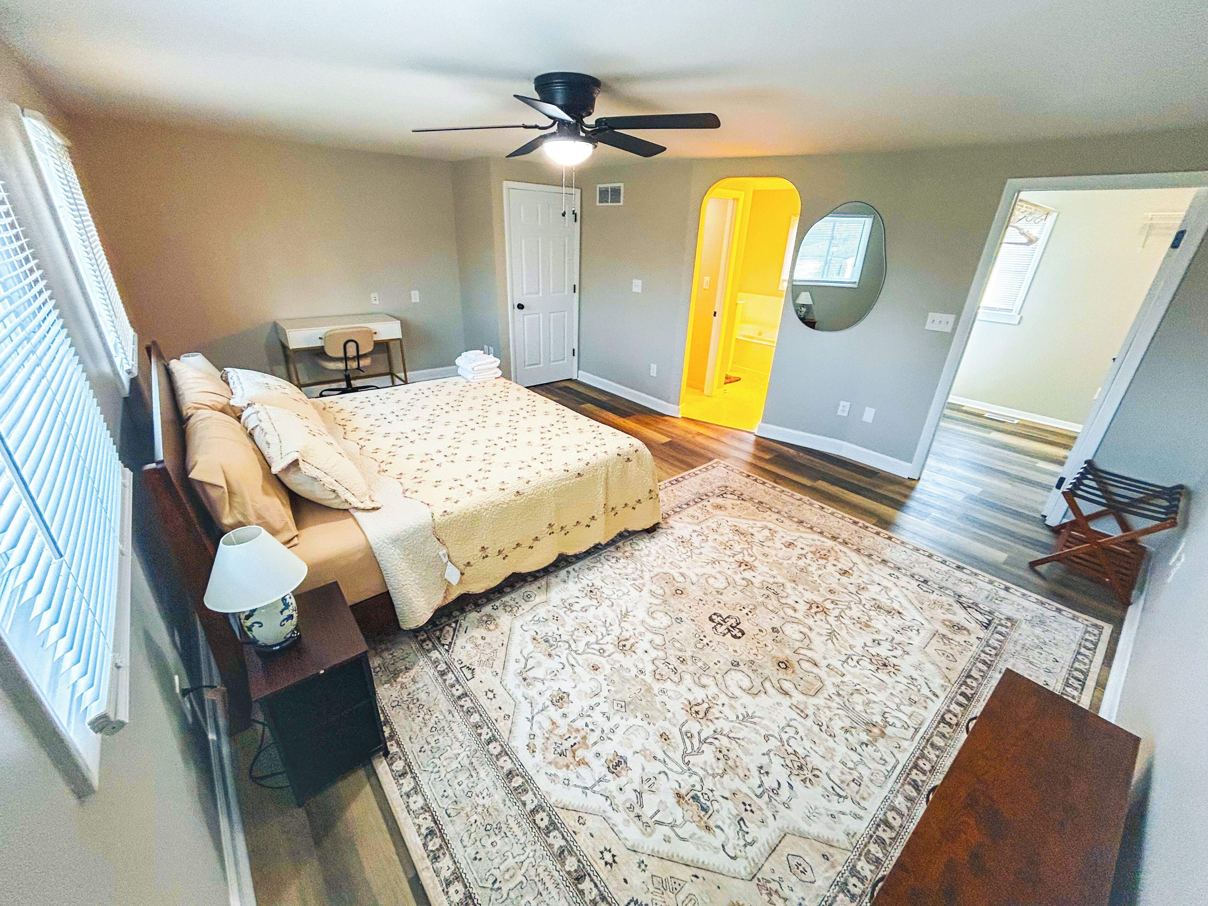 A bedroom with a bed, a desk, a chair, a ceiling fan, and a large rug.