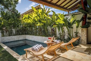 Zona junto a la piscina con sillones de descanso, una mesa con una bebida y exuberante follaje tropical.