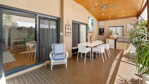 Um interior moderno com um deck de madeira, uma área de jantar com mobiliário branco e uma cozinha com geladeira e pia.