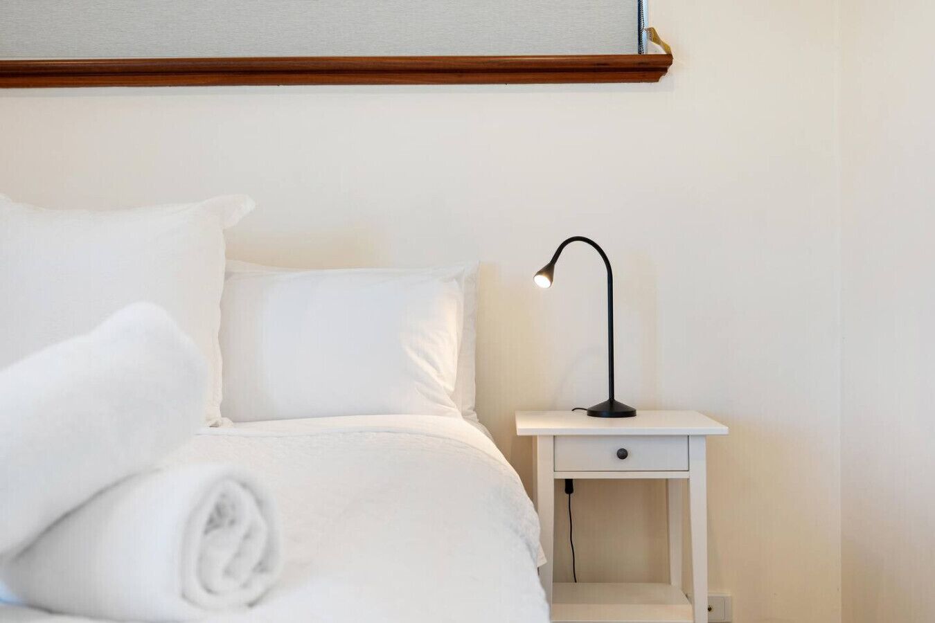 A neatly made bed with white linens and pillows, a small bedside table with a lamp, and a wooden headboard.