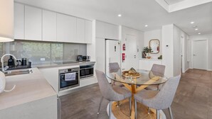 A modern kitchen with white cabinets, a glass dining table, and four grey chairs. - South Perth Sky - Foreshore Escape (Perth)