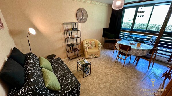A living room with a sofa, a small table, a bookshelf, a chair, and a dining area with a table and chairs. - Le Pradet A 781 - Mont Dore 4 Pers (Mont-Dore)