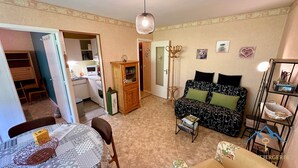 A cozy living room with a sofa, a dining table with chairs, a kitchen area, and a small bookshelf. - Le Pradet A 781 - Mont Dore 4 Pers (Mont-Dore)