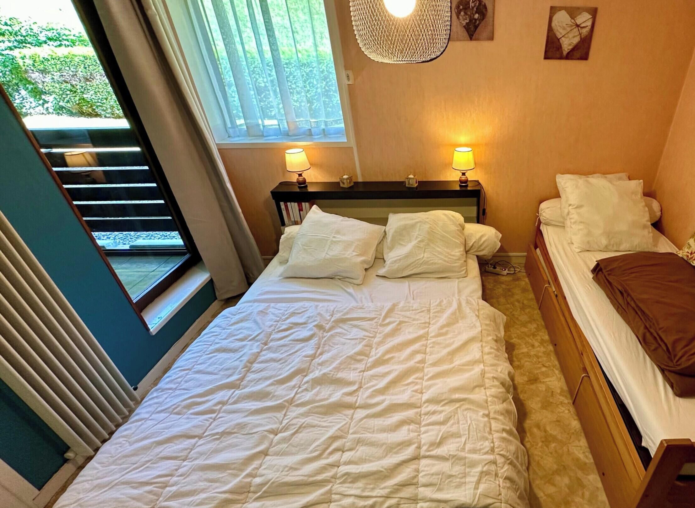 A bedroom with a bed, two bedside tables with lamps, a window with curtains, and a view of greenery outside.