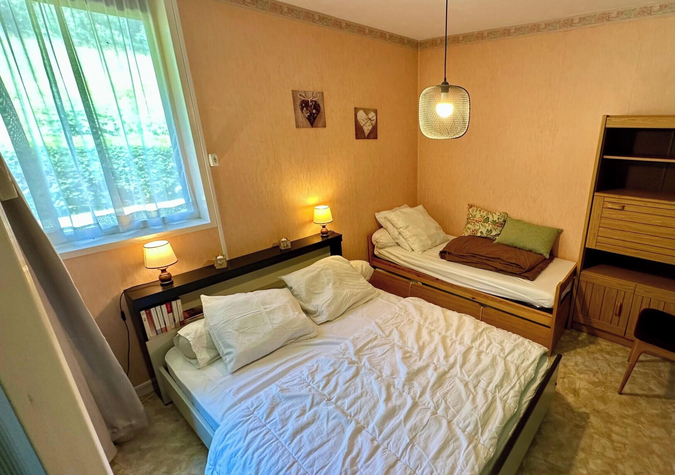 Et soveværelse med to senge, et vindue med gardiner, en senget lampe, en lille hylde og et klædeskab.