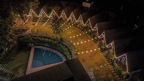 A pool surrounded by a garden with a series of buildings, each with a pointed roof and illuminated by string lights. - The Jungle Walker Resort  (Rishikesh)