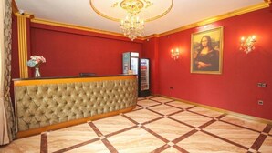 Un hall d’hôtel avec un mur rouge, un comptoir d’accueil capitonné, un lustre et une œuvre d’art encadrée.