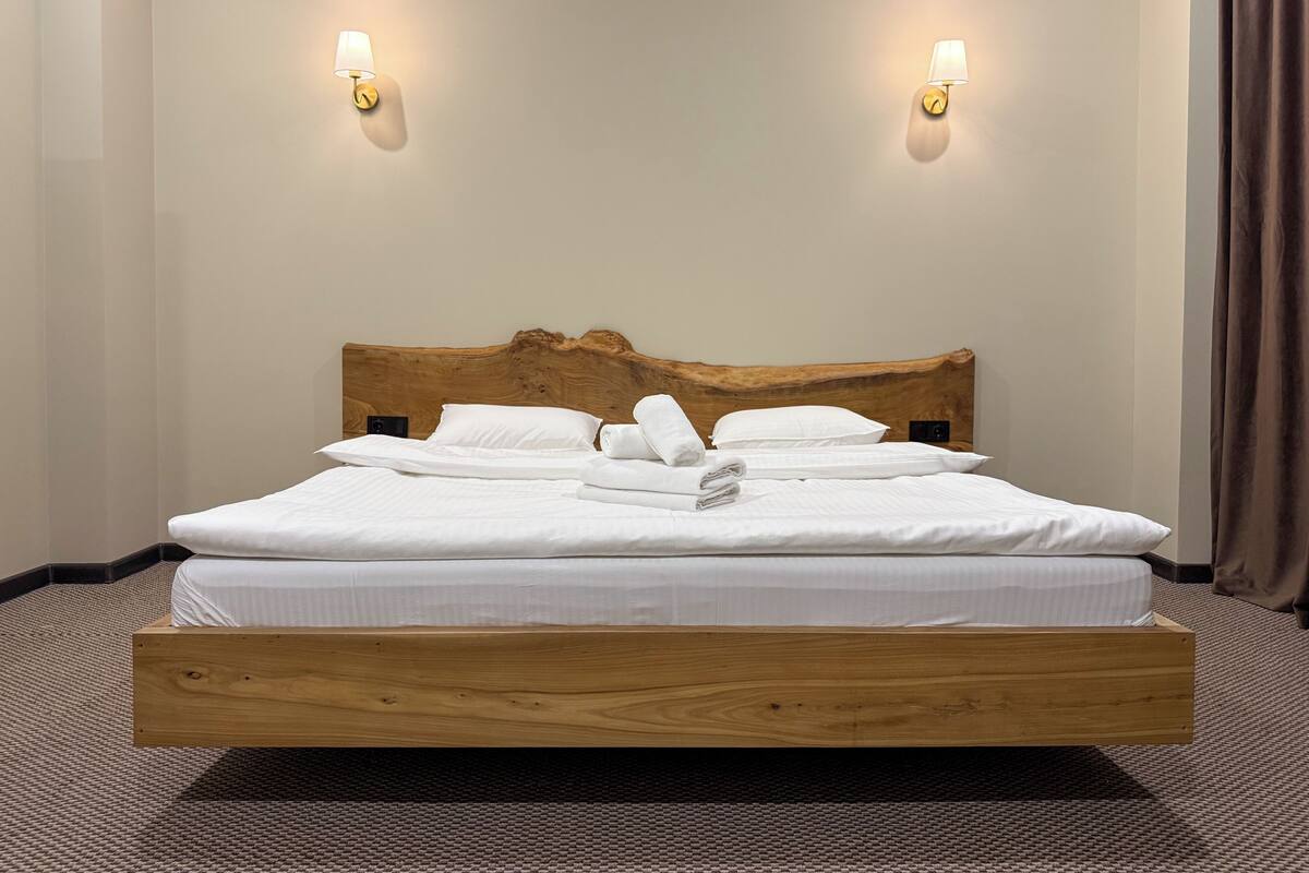 Una cama bien hecha con sábanas y toallas blancas, un cabecero de madera y dos lámparas fijadas a la pared.