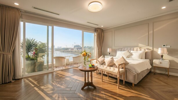 Una habitación de hotel con un ventanal grande, una cama, un sofá, una mesita con un florero y una mesita de noche con una lámpara.