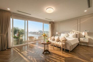 Una habitación de hotel con un ventanal grande, una cama, un sofá, una mesita con un florero y una mesita de noche con una lámpara.