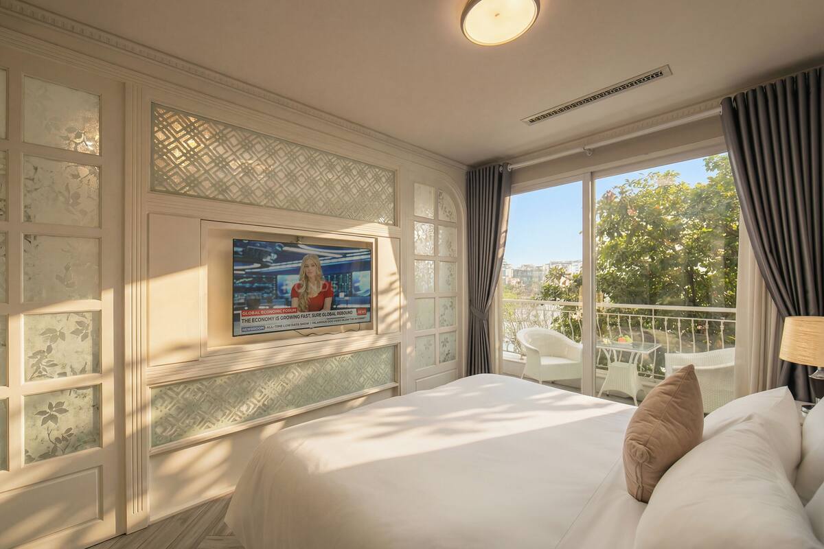 Una habitación de hotel con una cama grande, un televisor montado en la pared, un balcón con mobiliario exterior y un tratamiento decorativo en la ventana.