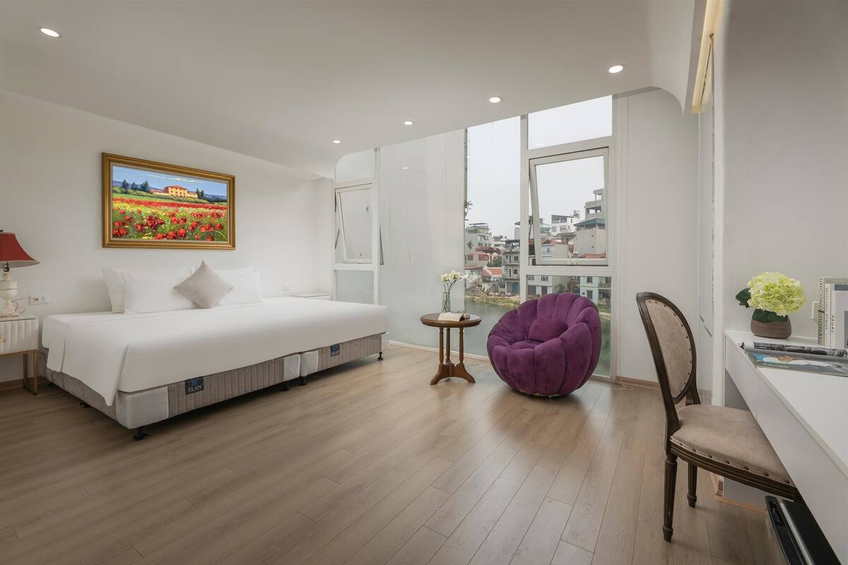 Una habitación de hotel moderna con una cama, una silla, una mesita y un ventanal grande que ofrece vistas a la ciudad.