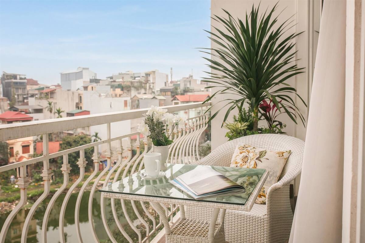 Un balcón con una mesa de vidrio, una silla de mimbre, una planta en maceta y vistas de edificios.