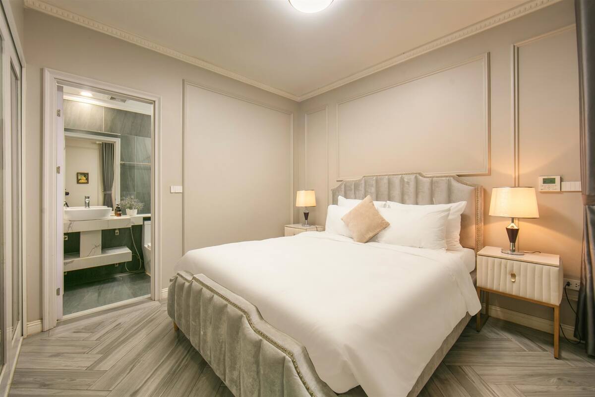 Habitación de hotel con una cama grande, dos mesitas de noche con lámparas, un espejo y un baño con lavabo y toallero.