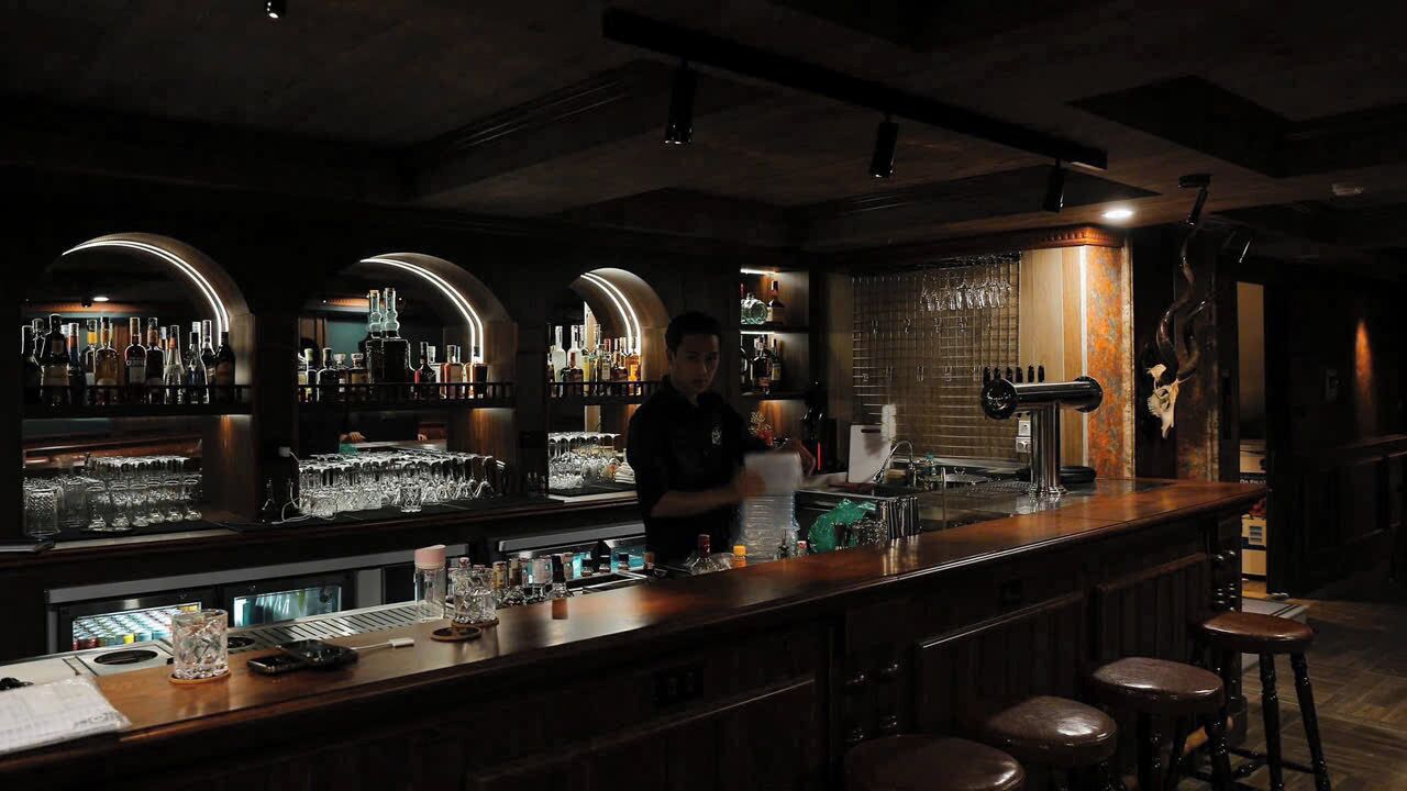 Un espace bar avec un barman, des étagères remplies de bouteilles, des verres et des tabourets.