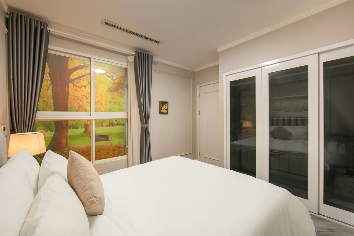 Une chambre d’hôtel avec un grand lit, une fenêtre avec des rideaux, une vue sur un parc et une porte-fenêtre coulissante donnant sur un balcon.