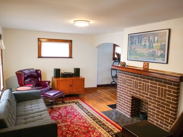 A living room with a fireplace, a red and gold patterned rug, a leather armchair, a sofa, and a wooden cabinet with speakers. - Family-Friendly Village Home - Downtown Location, 2 Car Garage, Big Backyard! (Columbus)