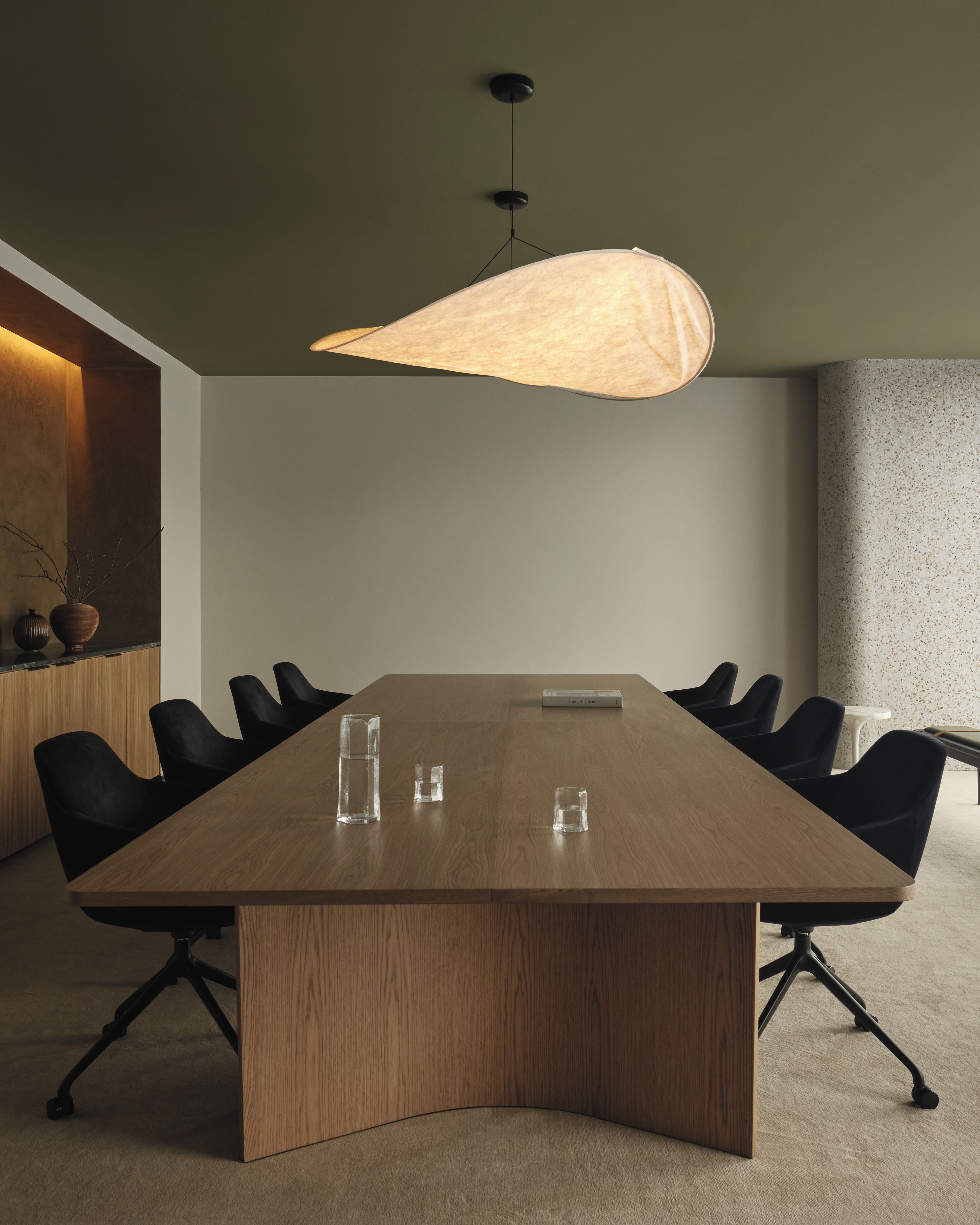 A conference room with a long table, black chairs, a hanging light fixture, and a vase on the sideboard.