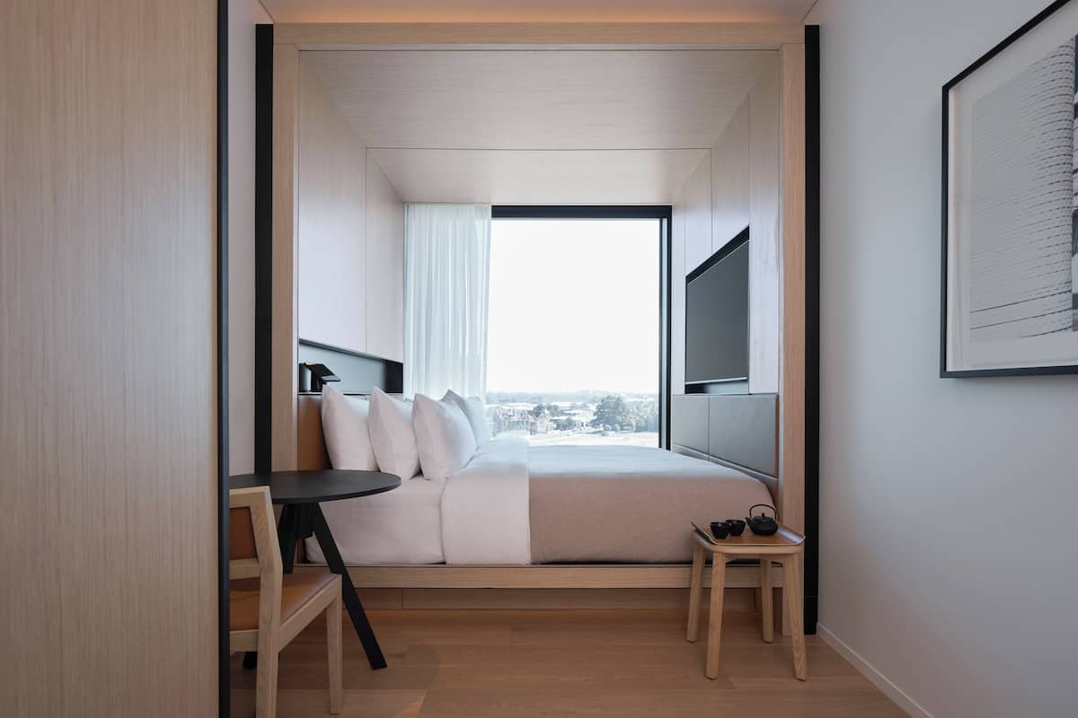 A modern hotel room with a bed, a small table, a TV mounted on the wall, and a large window offering a city view.