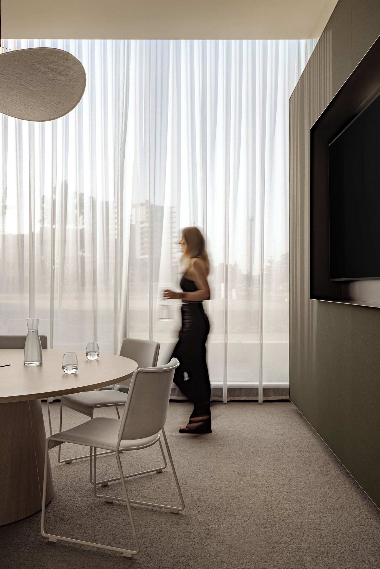 A modern hotel room with a large window, a round table with chairs, a flat-screen TV mounted on the wall, and a large pendant light.
