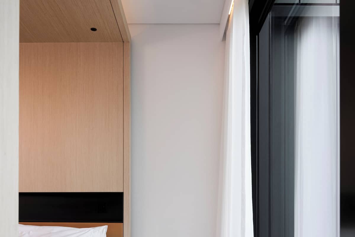 A modern hotel room with a bed, a small wooden stool, a window with curtains, and a built-in wardrobe.