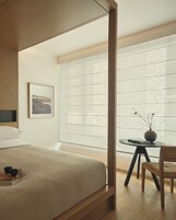 Un dormitorio con una cama, una mesita de noche con un florero, una silla y una ventana con persianas.