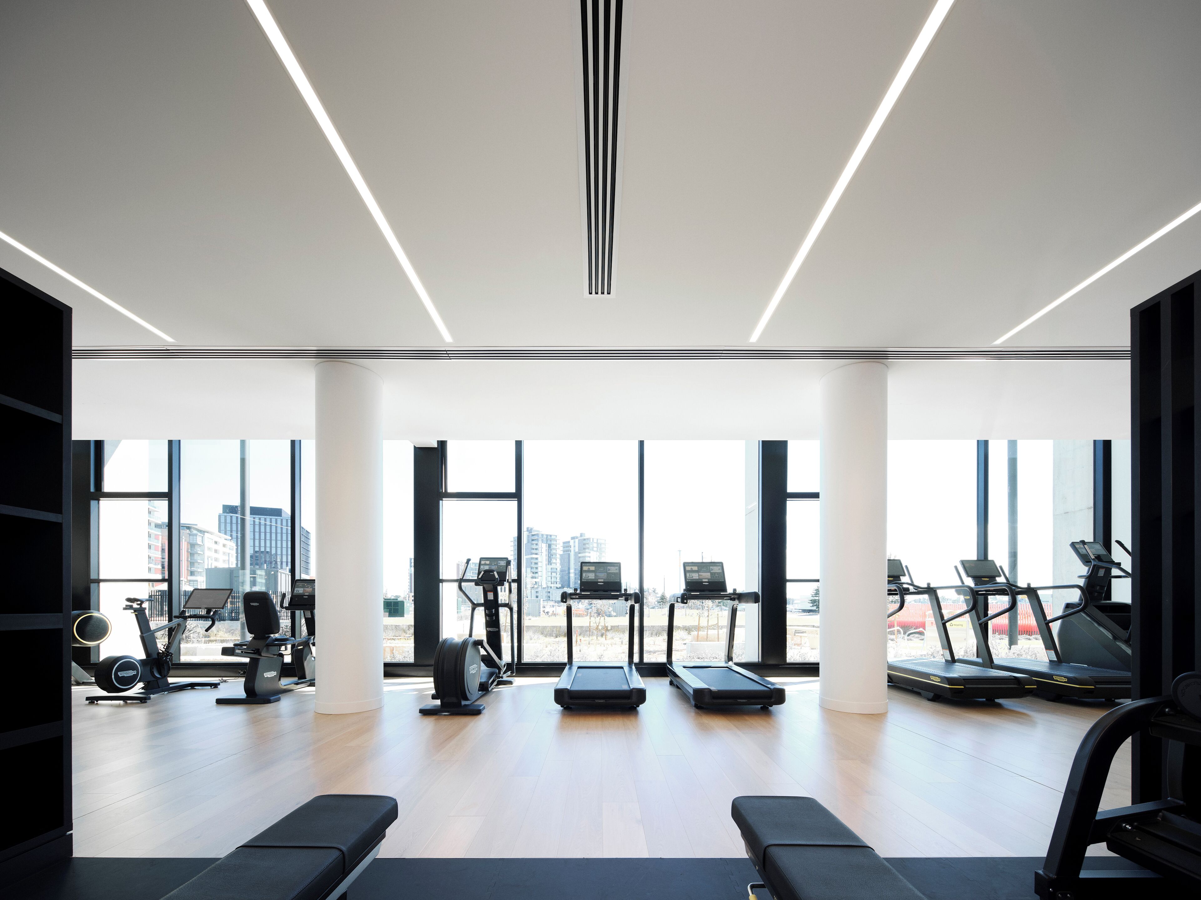 Un gimnasio moderno con amplios ventanales, diversos equipos de ejercicio y vistas a la ciudad.