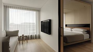 A modern hotel room with a sofa, a small table, a bed with a headboard, a TV mounted on the wall, and a large window with sheer curtains.