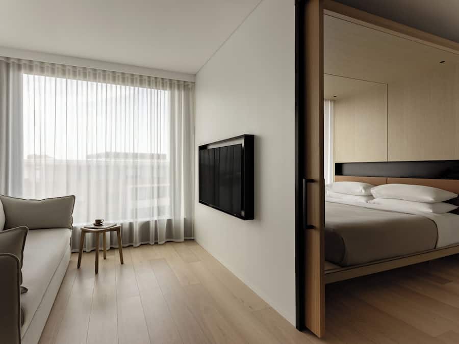 Una habitación de hotel moderna con sofá, una mesita, cama con cabecera, televisor montado en la pared y un ventanal con cortinas de gasa.