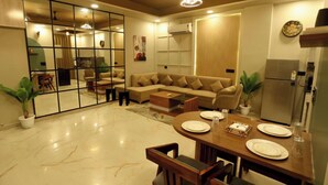 A modern living room with a sofa, dining table, and chairs. There is a kitchen area with a refrigerator and a clock on the wall. - Motwani Stays By Dr Richa Motwani (Jaipur)