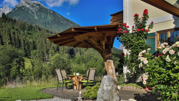 A patio with a wooden table and chairs, surrounded by a garden with flowers and a small pond. - Grünstein - 2-room apartment, south and east-facing balcony, 2 persons, 40 sqm, safe, kitchenette (Ramsau bei Berchtesgaden)