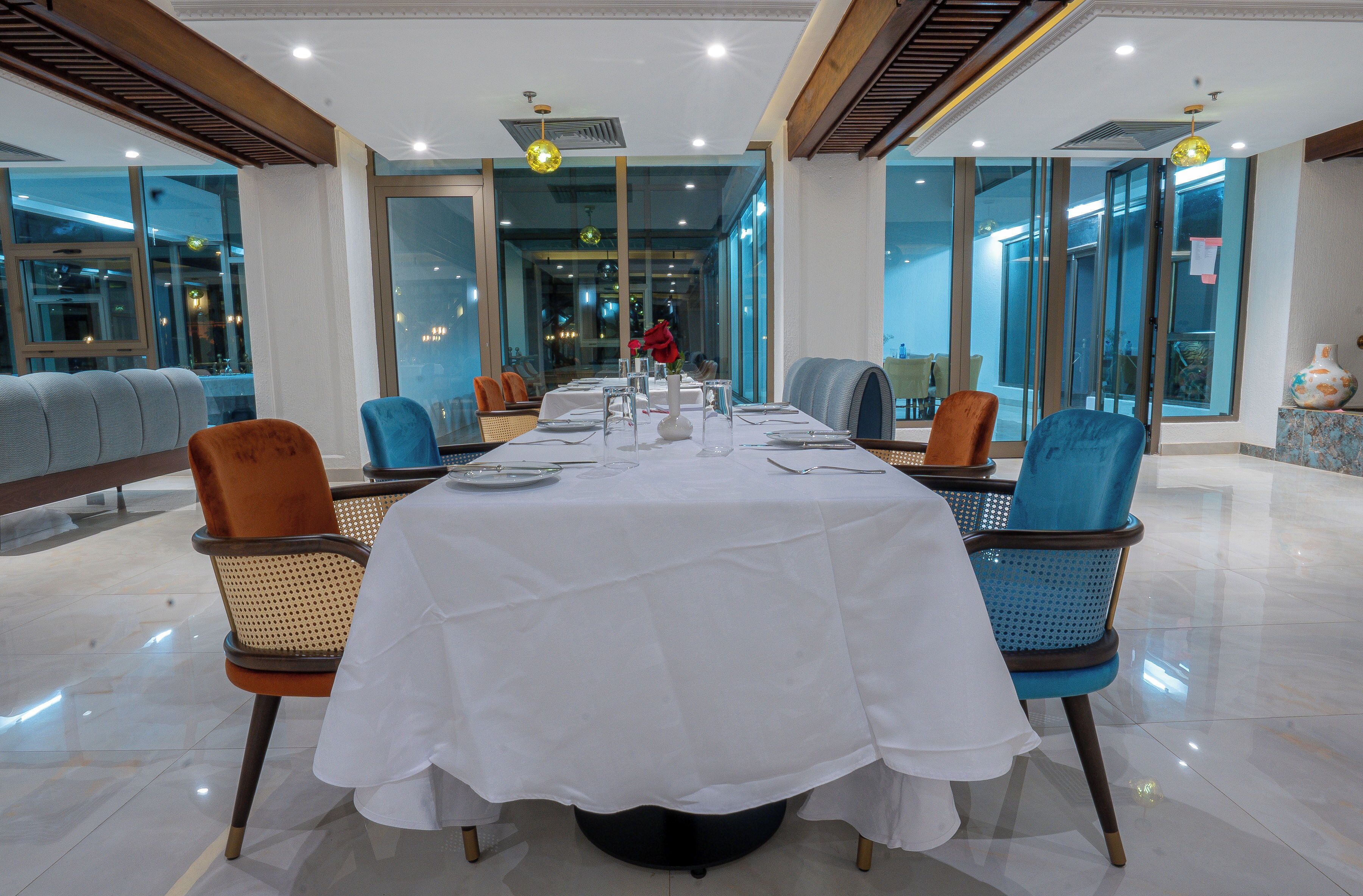 Un comedor con una mesa larga dispuesta para una comida, rodeada de sillas y grandes ventanales que ofrecen vistas del interior.